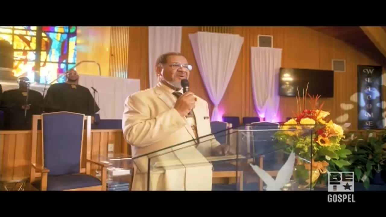 A man in a cream-colored suit speaks into a microphone, standing behind a clear podium in a church. Behind him, a choir stands on a raised platform, and a stained-glass window glows in the background.
