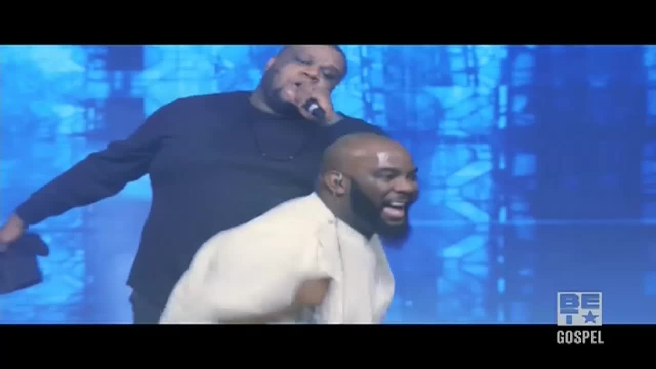 On a BET Gospel stage, a man in a black shirt sings into a microphone, while another man in white, beside him, throws his arms in the air, eyes wide with joy. The background is a wash of blue light.
