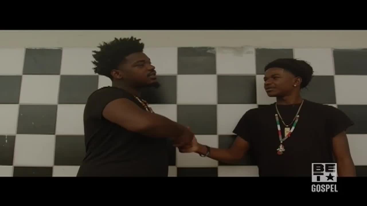 Two young men, both wearing black t-shirts, clasp hands in a firm handshake. The backdrop is a simple checkerboard pattern, and the BET Gospel logo is visible in the corner.
