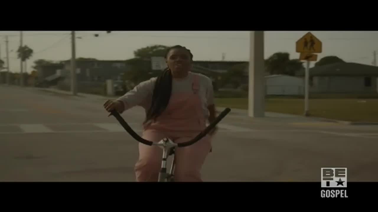 A woman in pink overalls pedals a bicycle down a residential street. The BET Gospel logo is in the corner, and the scene has a warm, hazy quality.
