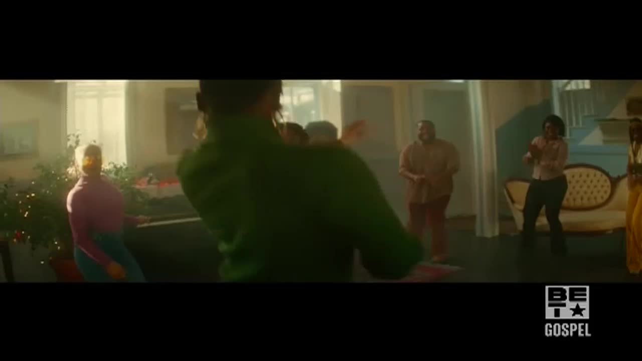 People are dancing in a bright, sunlit room. A man in a green shirt leads the movement, while others clap and sway to the music.
