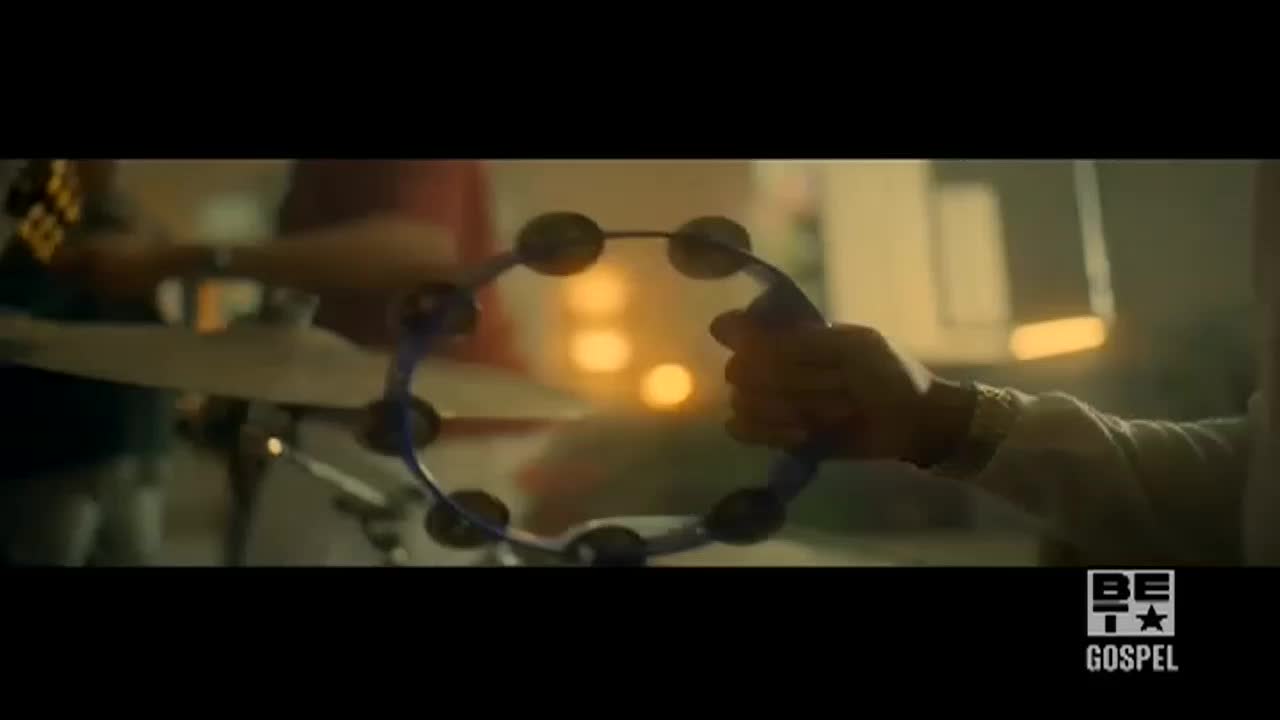 A hand holds a blue tambourine, the jingles ready to ring out. In the background, a drummer's arm moves, and the scene glows with warm light.
