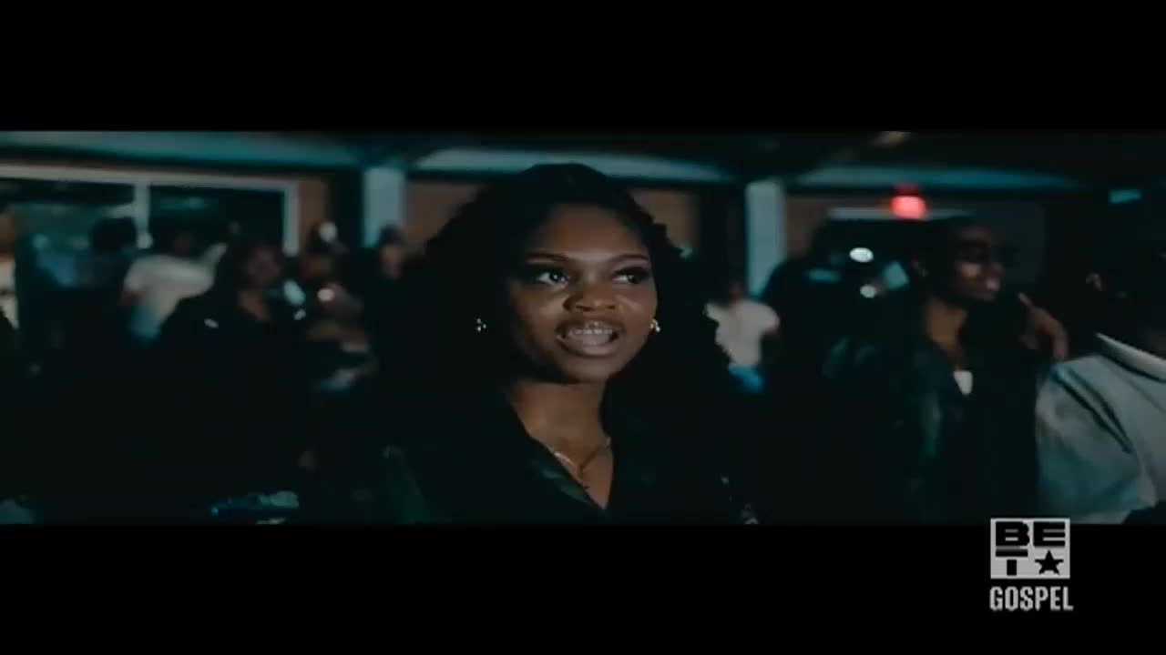 A young woman with curly hair is speaking, her expression animated.  Behind her, a crowd of people are gathered in a dimly lit space, likely at a BET Gospel event.
