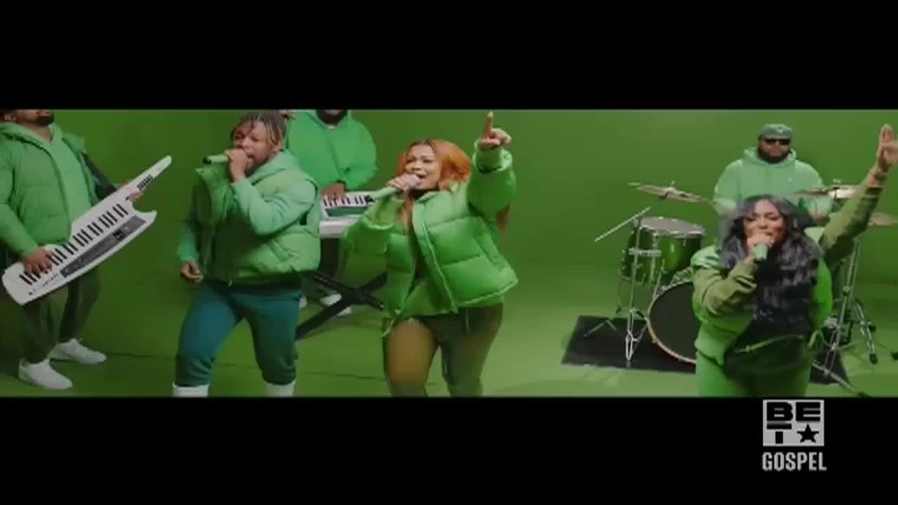 A gospel band, all in matching green, is performing on a green screen stage. The lead singers are gesturing with their hands, microphones in hand, while the band plays.
