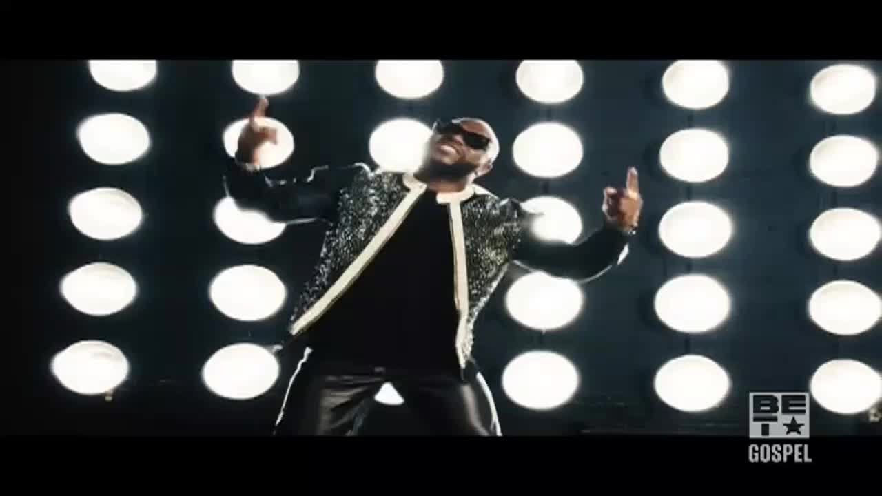 A man in a sparkly jacket and shades gestures with both hands, his fingers pointing upwards. Behind him, a wall of bright, round lights illuminates the stage.
