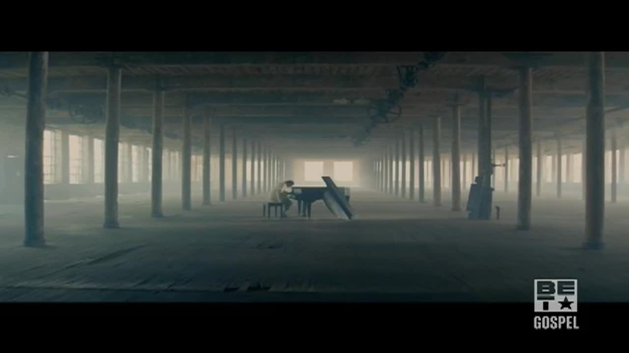 A musician sits at a grand piano, playing in a cavernous, empty space. The scene feels like a performance on BET Gospel, with the piano's dark shape contrasting with the light at the end of the long, industrial hall.
