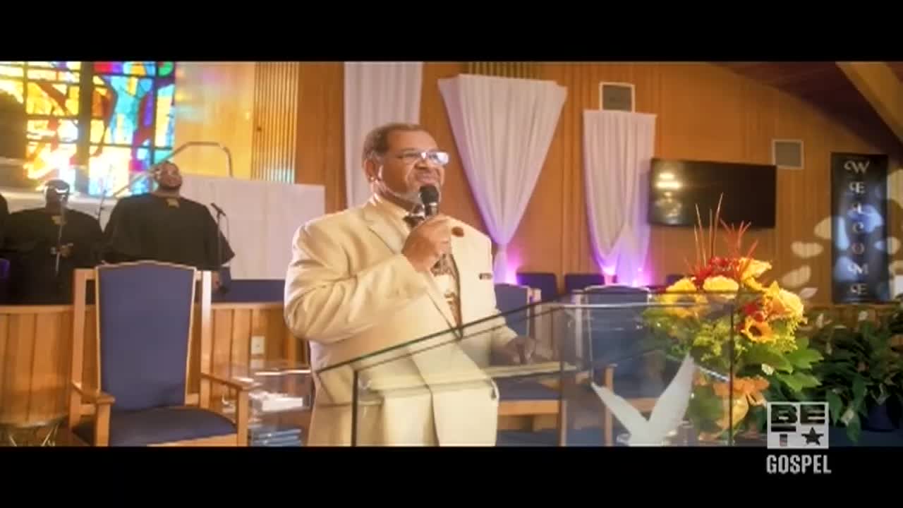 A man in a cream suit stands at a podium, speaking into a microphone. Behind him, a choir in black robes stands ready on the stage in a church, as the BET Gospel logo sits in the corner.
