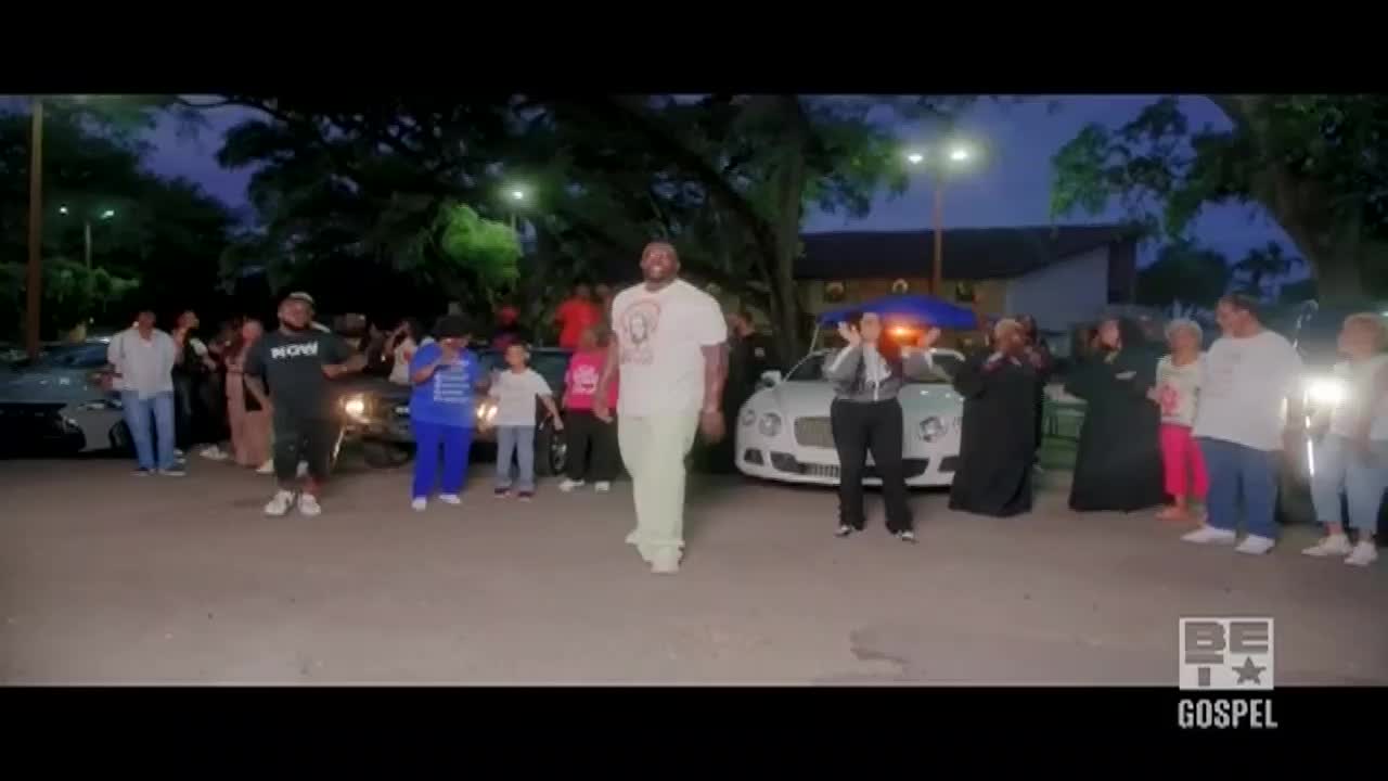 A man in a white tee and light pants walks toward the camera as a crowd cheers. Behind him, a Bentley sits parked, and the BET Gospel logo is in the corner.
