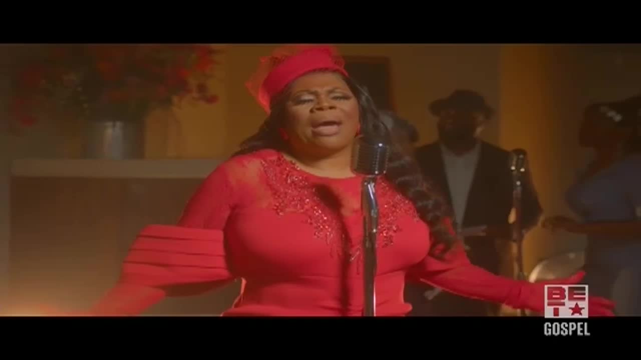 A woman in a red dress and hat belts into a vintage microphone, her arms outstretched. Behind her, a band provides accompaniment on a dimly lit stage.
