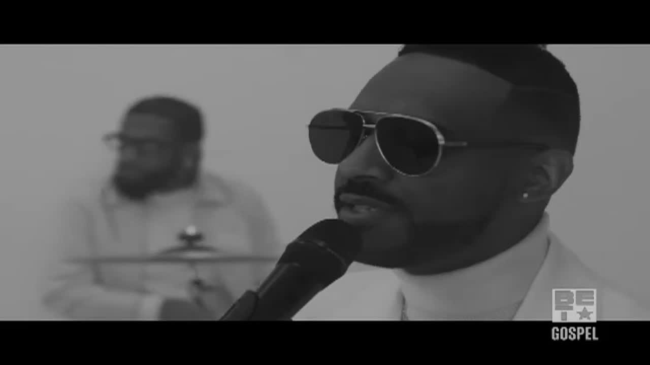 A man in sunglasses sings into a microphone, his face lit in the dim light. Behind him, another man is slightly out of focus, playing drums.
