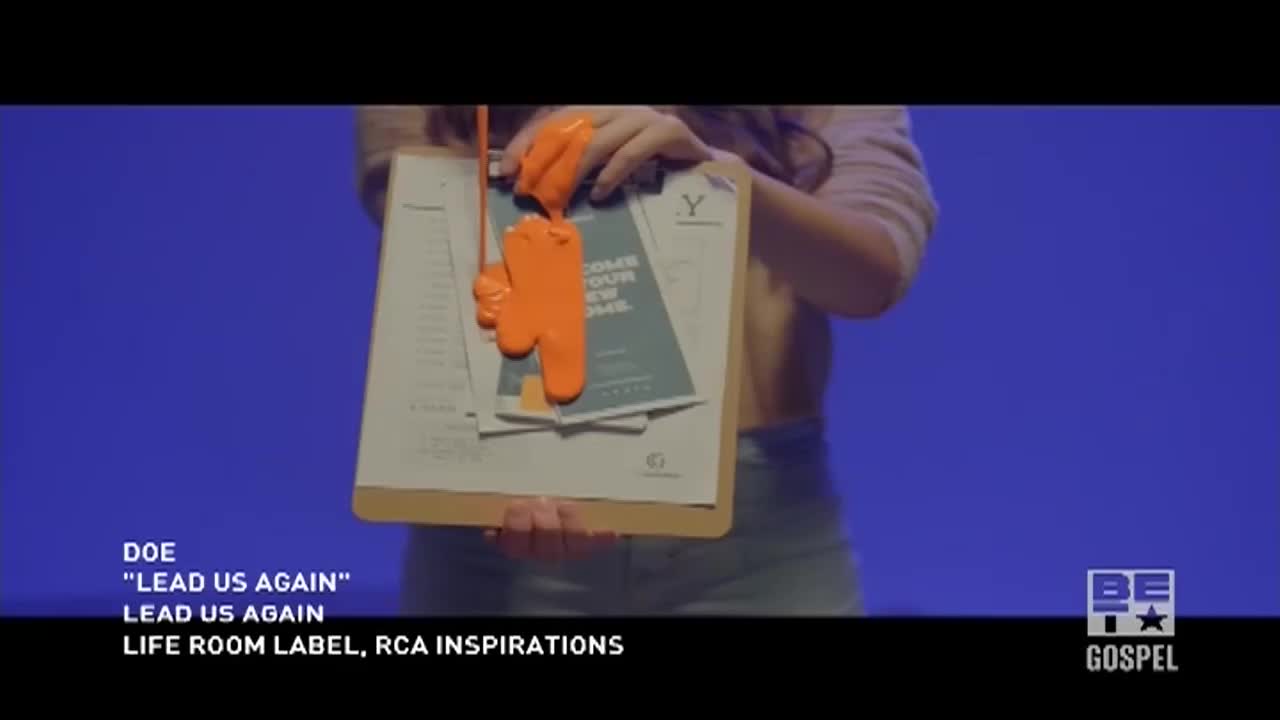 A woman holds a clipboard with papers, and a bright orange substance drips down onto them. It's a scene from a BET Gospel music video, with the words "Lead Us Again" visible.
