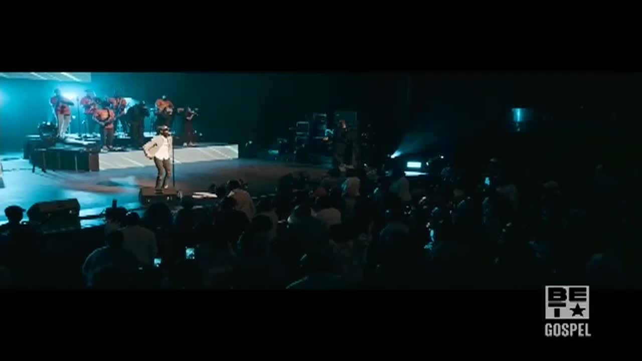 A singer in a white shirt and dark pants stands on a small platform, microphone in hand. The stage is lit with a cool blue light, and the crowd is packed in front of the stage.
