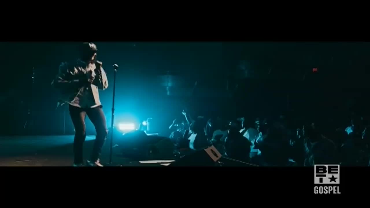A performer stands center stage, bathed in blue light, holding a microphone. The audience in the background has their hands raised, clearly enjoying the BET Gospel concert.
