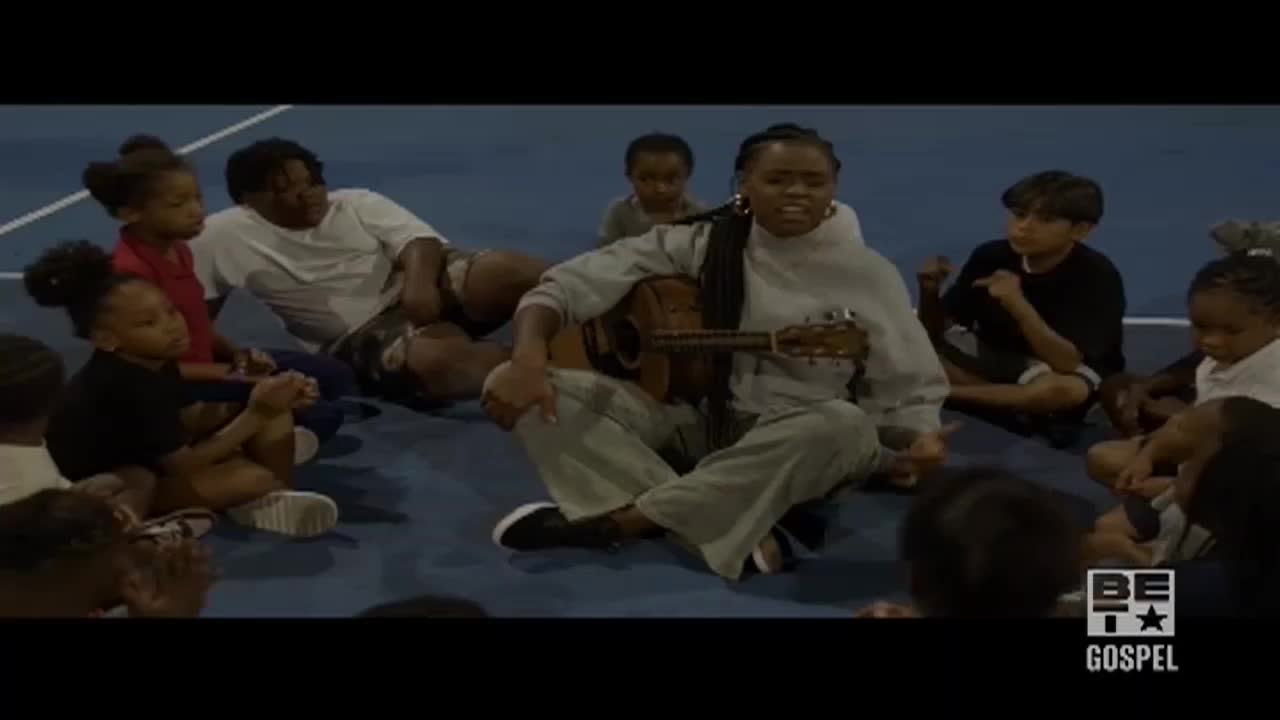 A woman sits cross-legged, strumming a small guitar while surrounded by children. They're all on a blue surface, and the BET Gospel logo sits in the corner.
