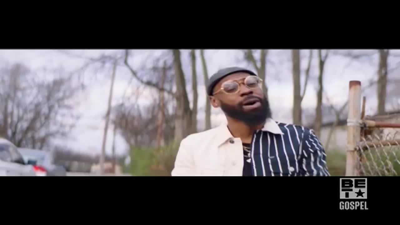 A man with a beard and glasses, wearing a hat and a jacket, is singing. He's standing outside, with bare trees and a chain-link fence behind him.
