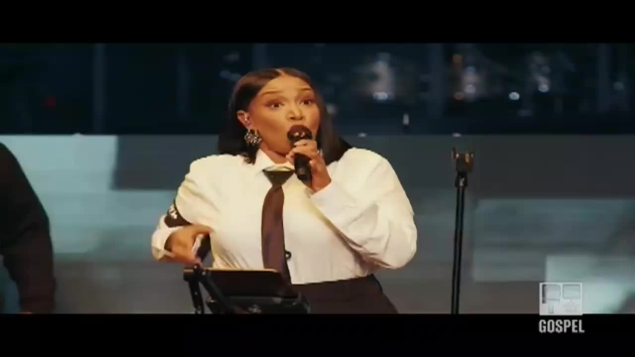 A woman in a white shirt and tie belts out a song, her mouth wide open as she holds a microphone. A "Gospel" logo is visible in the corner, and the stage is dimly lit.
