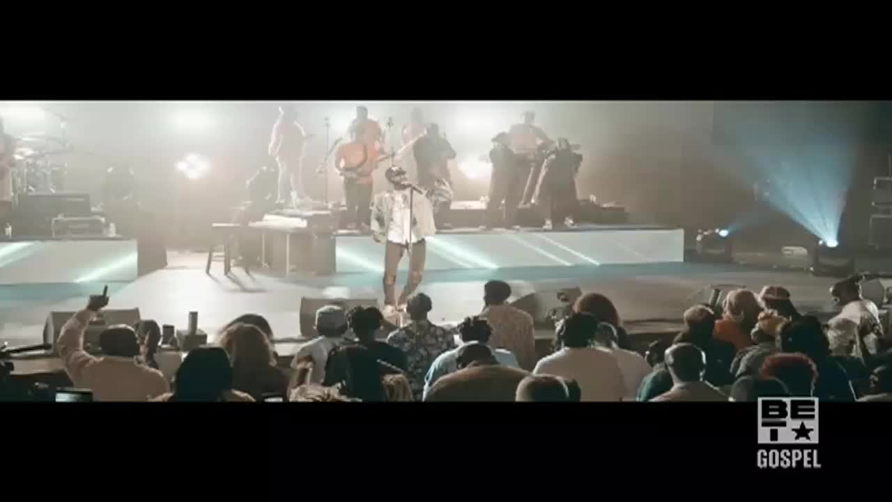 A singer in a white shirt stands center stage, microphone in hand, with a band behind him. The audience in front of the stage is lit by stage lights, and some people are raising their hands.
