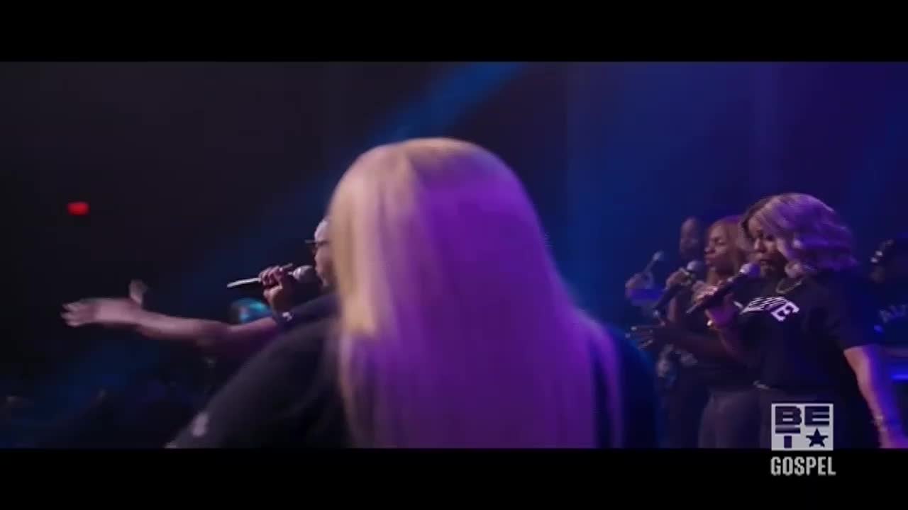 A woman with long, blonde hair is centered, her back to the camera, as a group of singers perform on stage. The BET Gospel logo is visible in the lower right corner.
