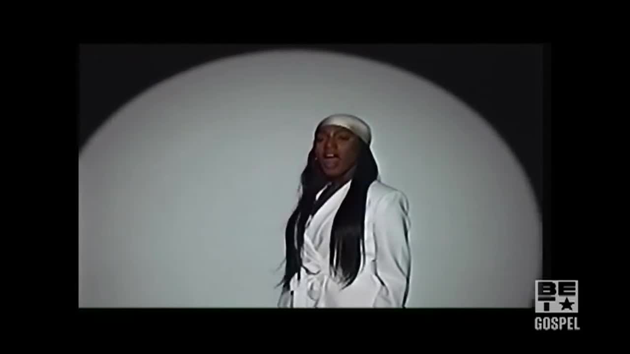 A woman in a white suit and headscarf stands in a spotlight, singing with her mouth open. The BET Gospel logo is visible in the corner.
