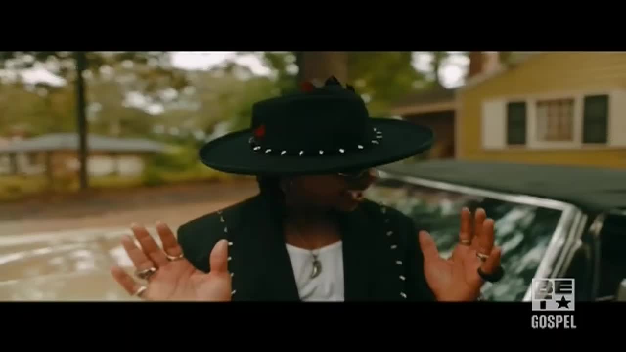A person in a studded hat and black jacket gestures with both hands, framed by a vintage car. The BET Gospel logo sits in the corner, and the scene appears to be filmed in a residential area in the United States.
