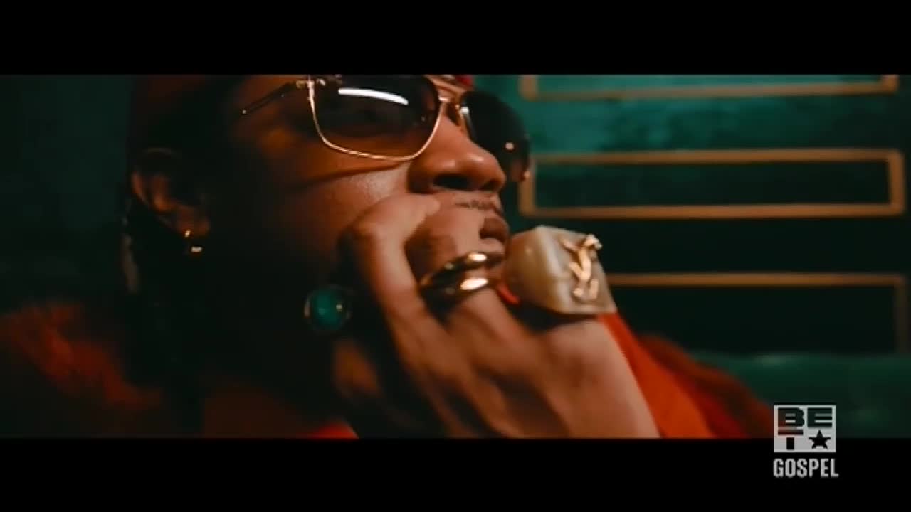 The man, wearing sunglasses and a red fur-trimmed jacket, rests his chin on his hand. His fingers are adorned with several rings, including a large one with a gold emblem.
