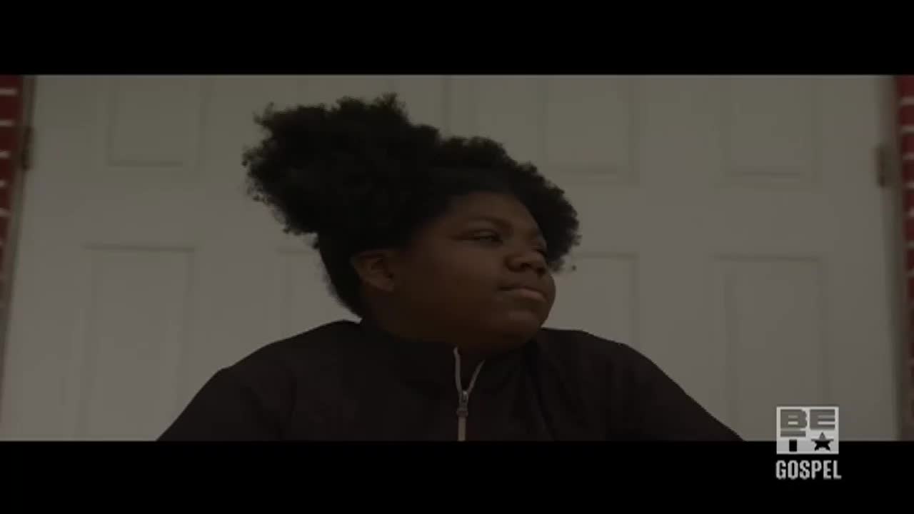A young person with a full afro stands in front of a white door, looking off to the side. The BET Gospel logo is visible in the corner.
