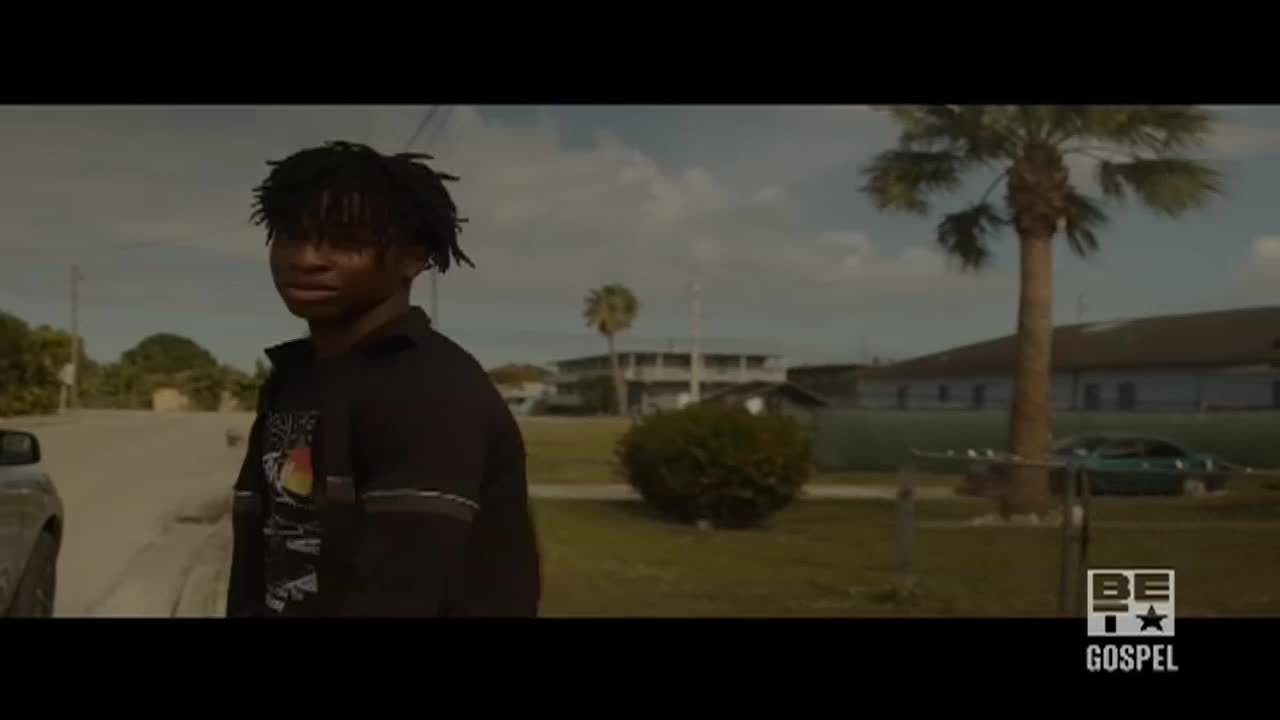 A young man in a black shirt stands on a street corner, looking toward the camera. The scene, set in a residential area, is likely part of a BET Gospel production.
