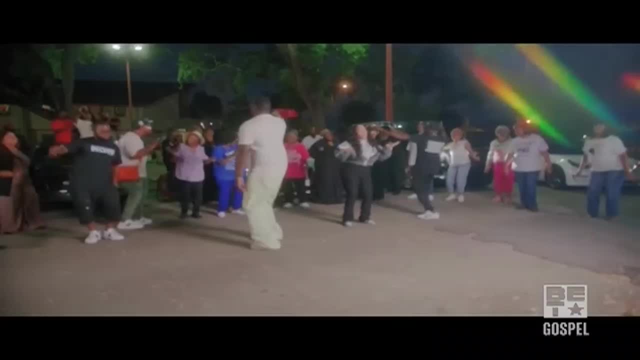 A crowd of people, illuminated by stage lights, are dancing in a parking lot. The scene, likely part of a BET Gospel event, shows folks swaying and clapping in time with the music.
