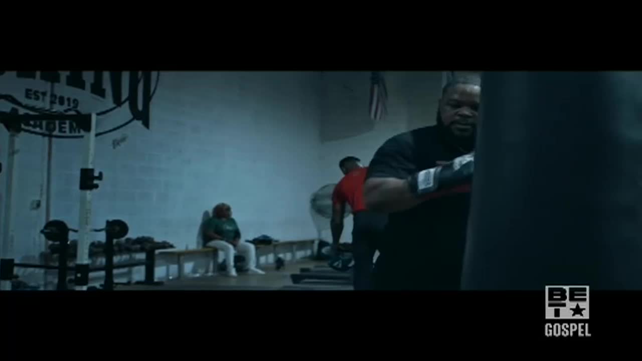A man in boxing gloves throws punches at a heavy bag, sweat beading on his forehead. In the background, a woman sits on a bench, watching the workout in the gym.
