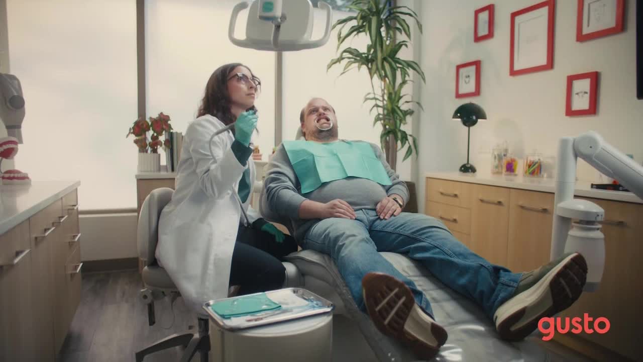 A dentist, wearing glasses and a lab coat, holds a dental drill poised over a patient's open mouth. The man reclines in the chair, a bib protecting his clothes as he stares up at the ceiling.
