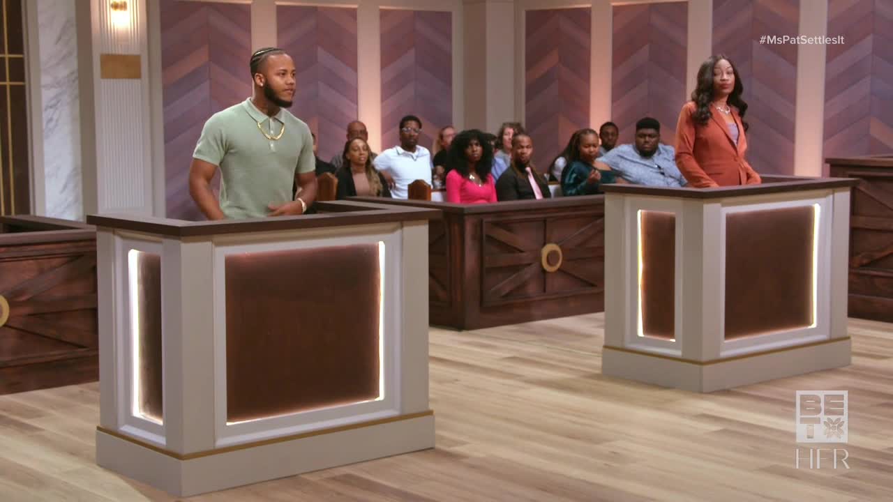 Two people stand at podiums, facing the audience, in what looks like a courtroom. A panel of people sits in the background, watching the proceedings.
