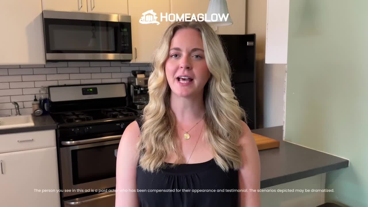 A woman with long blonde hair is standing in a kitchen, talking directly to the camera. Behind her are stainless steel appliances and a countertop.
