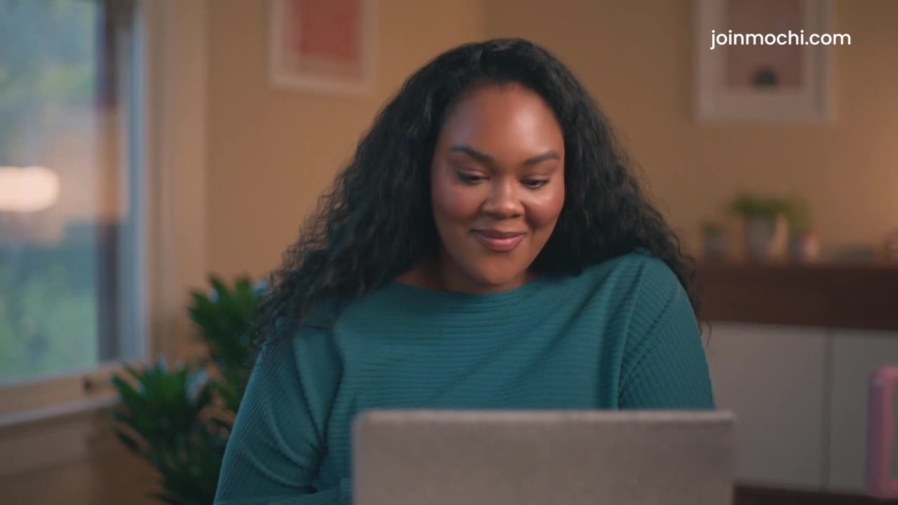 A woman with long, dark hair smiles at a laptop screen. The soft light from a window illuminates her face as she works.
