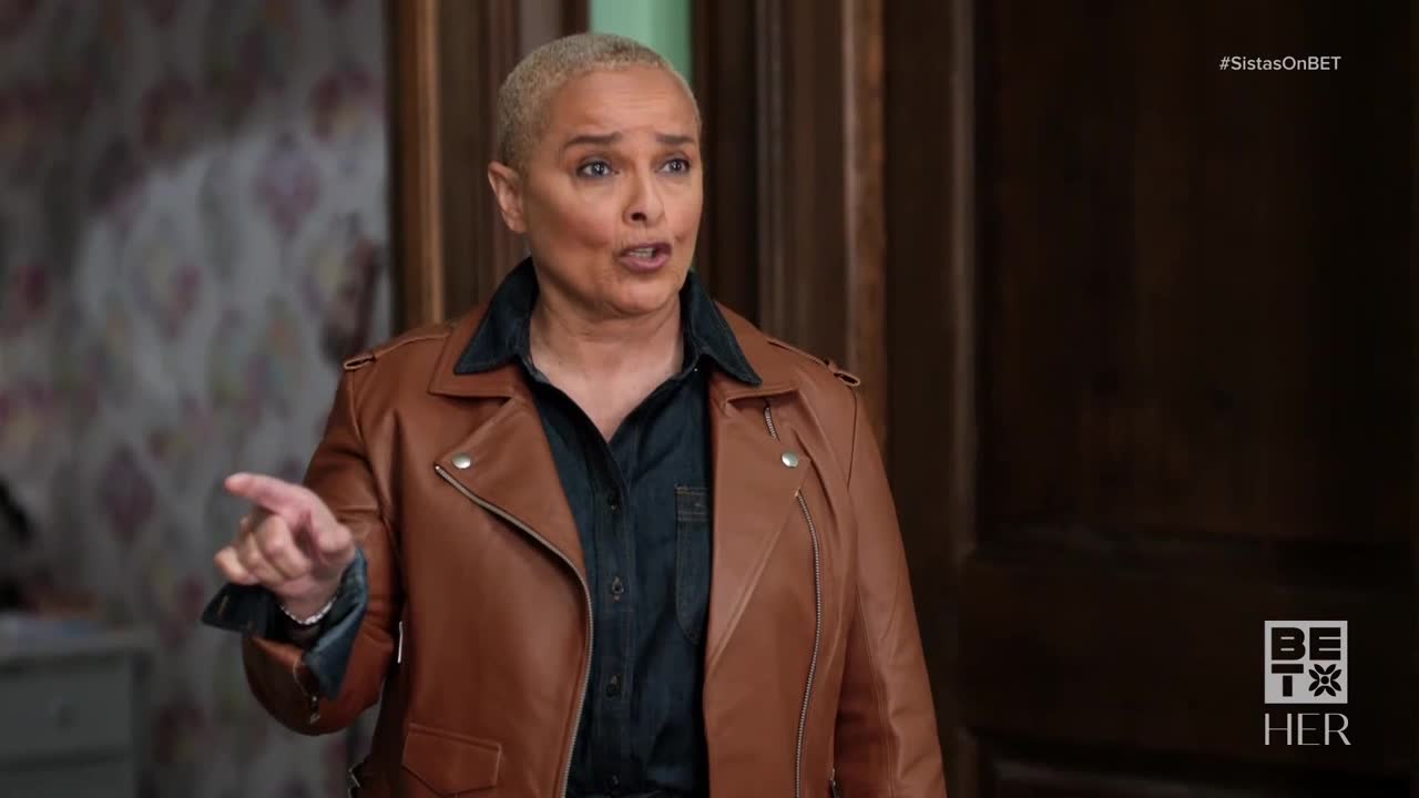 She's gesturing with her right hand, index finger extended, as she speaks. The woman is wearing a brown leather jacket over a dark denim shirt.
