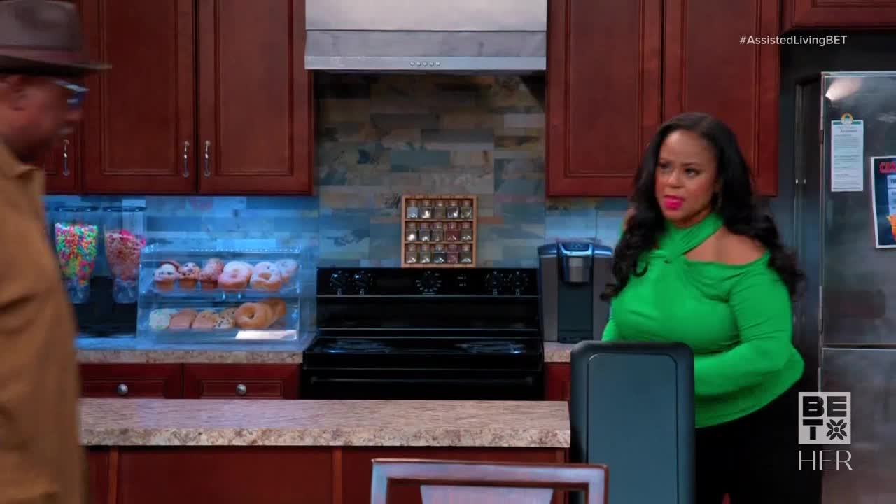 A woman in a bright green top stands in a kitchen, looking to her left. A man in a brown hat is walking toward her.
