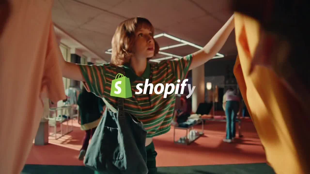 A young woman, wearing a striped shirt, holds up clothing racks with both arms. Behind her, people browse in a brightly lit space.
