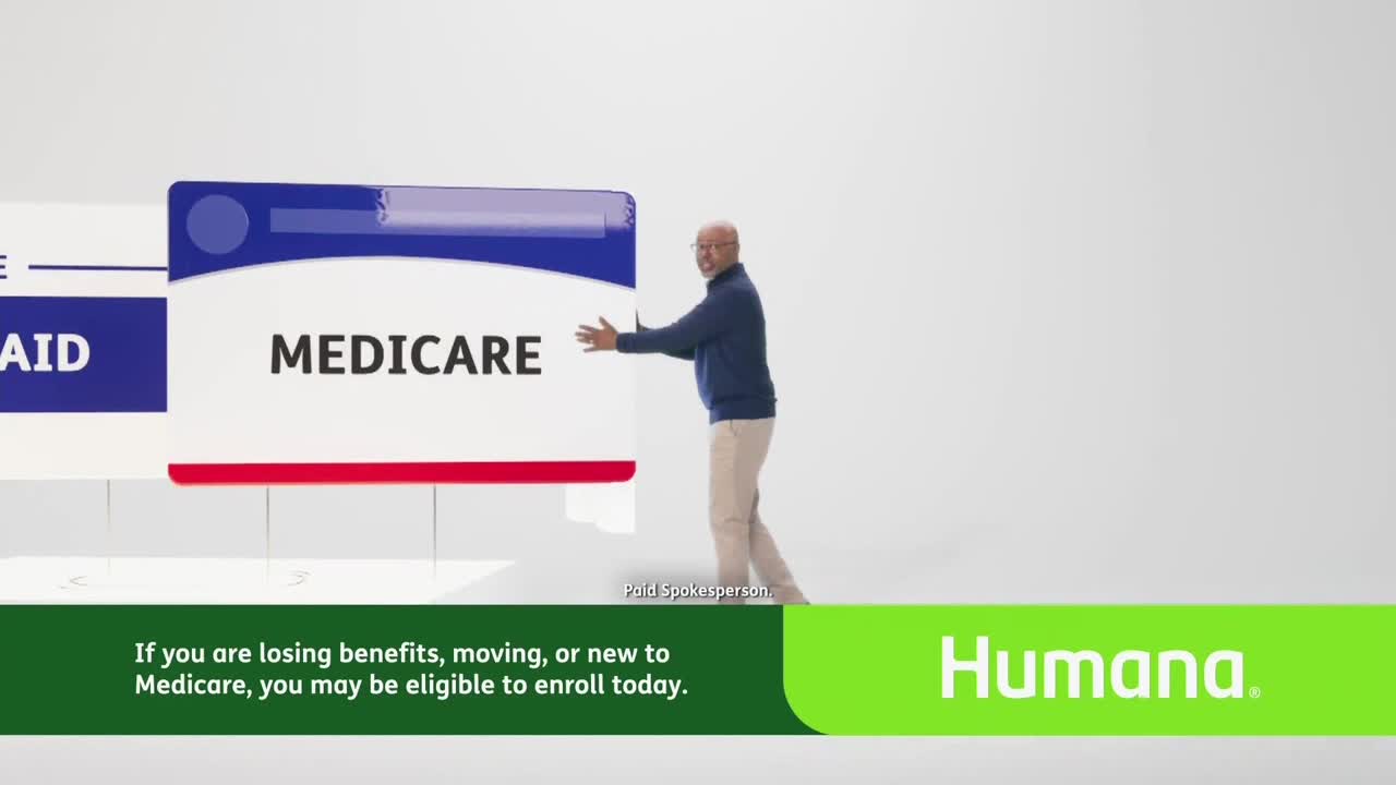 A man in a blue sweater pushes a large, rectangular sign that says "MEDICARE." The sign is white with a red stripe and a blue top.
