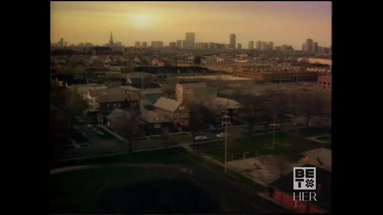 The sun sets over a city skyline, casting a warm glow on the rooftops. Below, the residential streets and a park are quiet, the day winding down.
