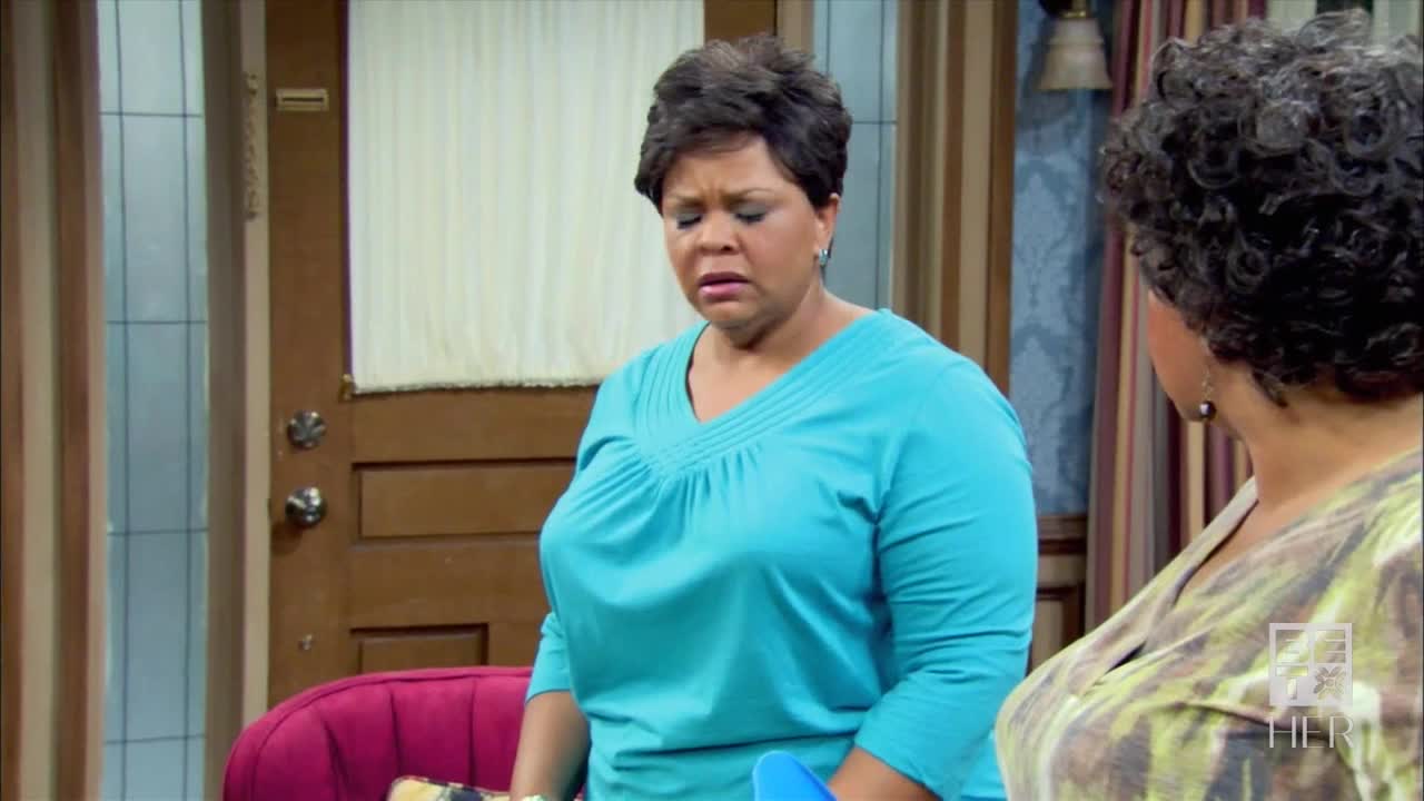 The woman in the blue shirt is frowning, her eyes downcast as she listens to the other woman. The scene unfolds in what looks like a living room, with a door and a curtain visible in the background.
