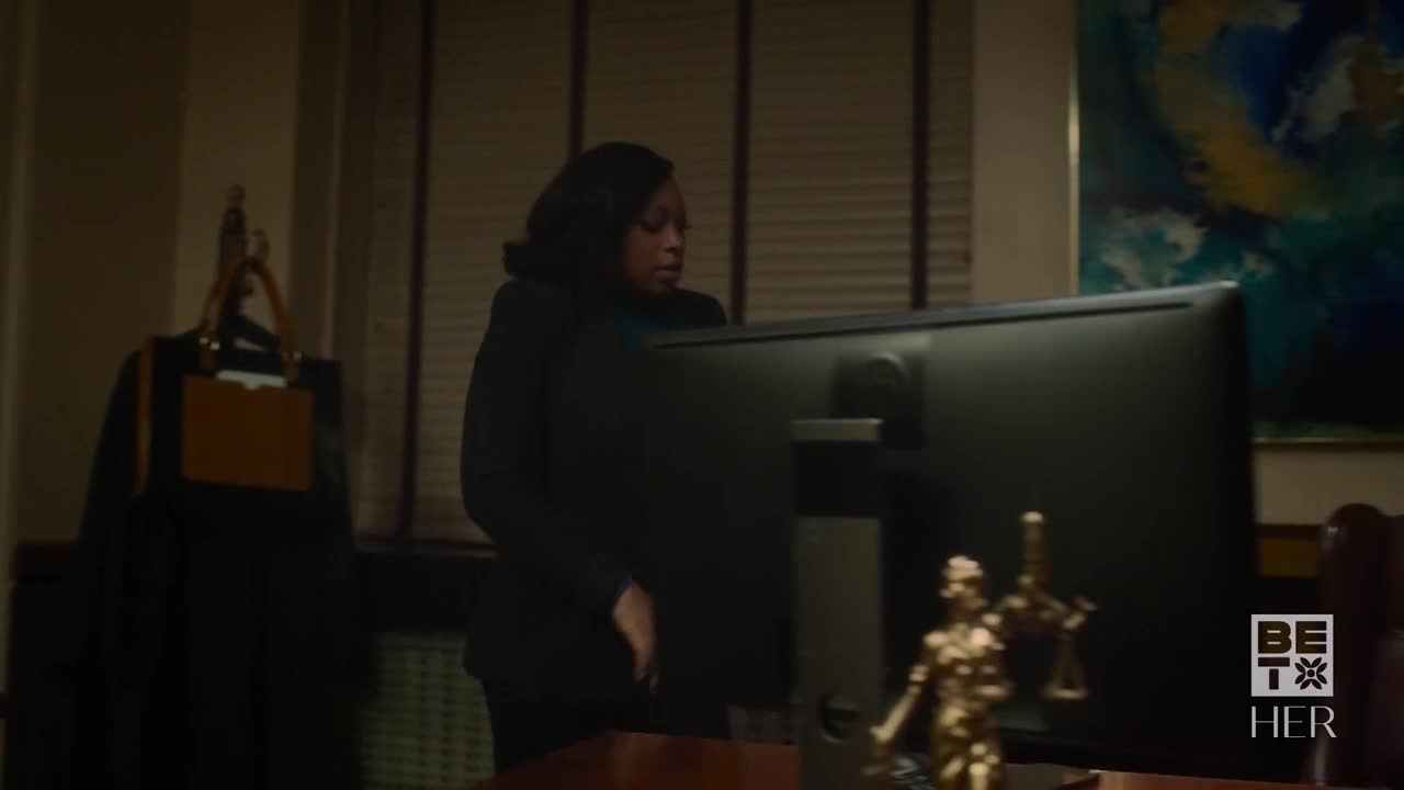 A woman in a dark suit stands in front of a large computer screen, her gaze fixed on it. A small statue of Lady Justice sits on the desk, and the BET Her logo is visible in the corner.
