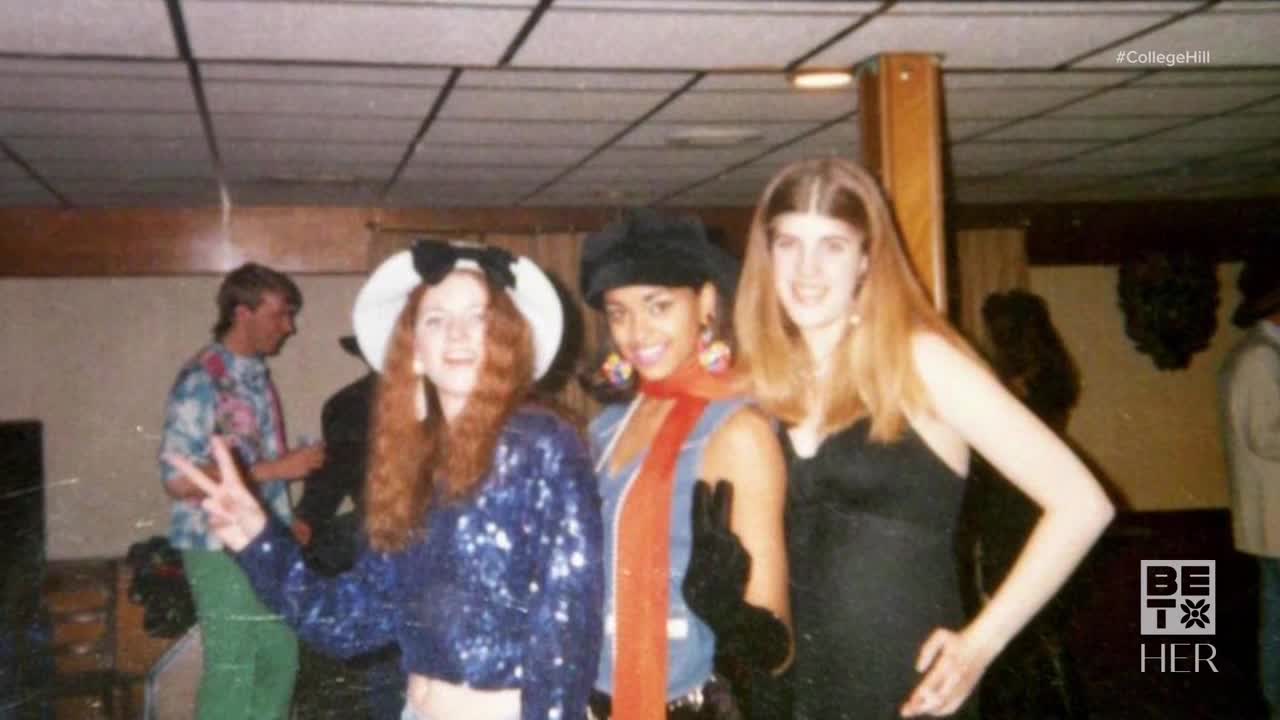 Three women pose together, smiling, with one flashing a peace sign. In the background, a man in a tie-dye shirt stands near a bar.
