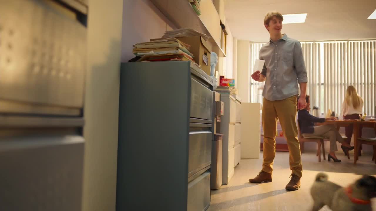 A young man, holding a stack of papers, stands in a bright office. A small dog trots past him, while people work at a table in the background.
