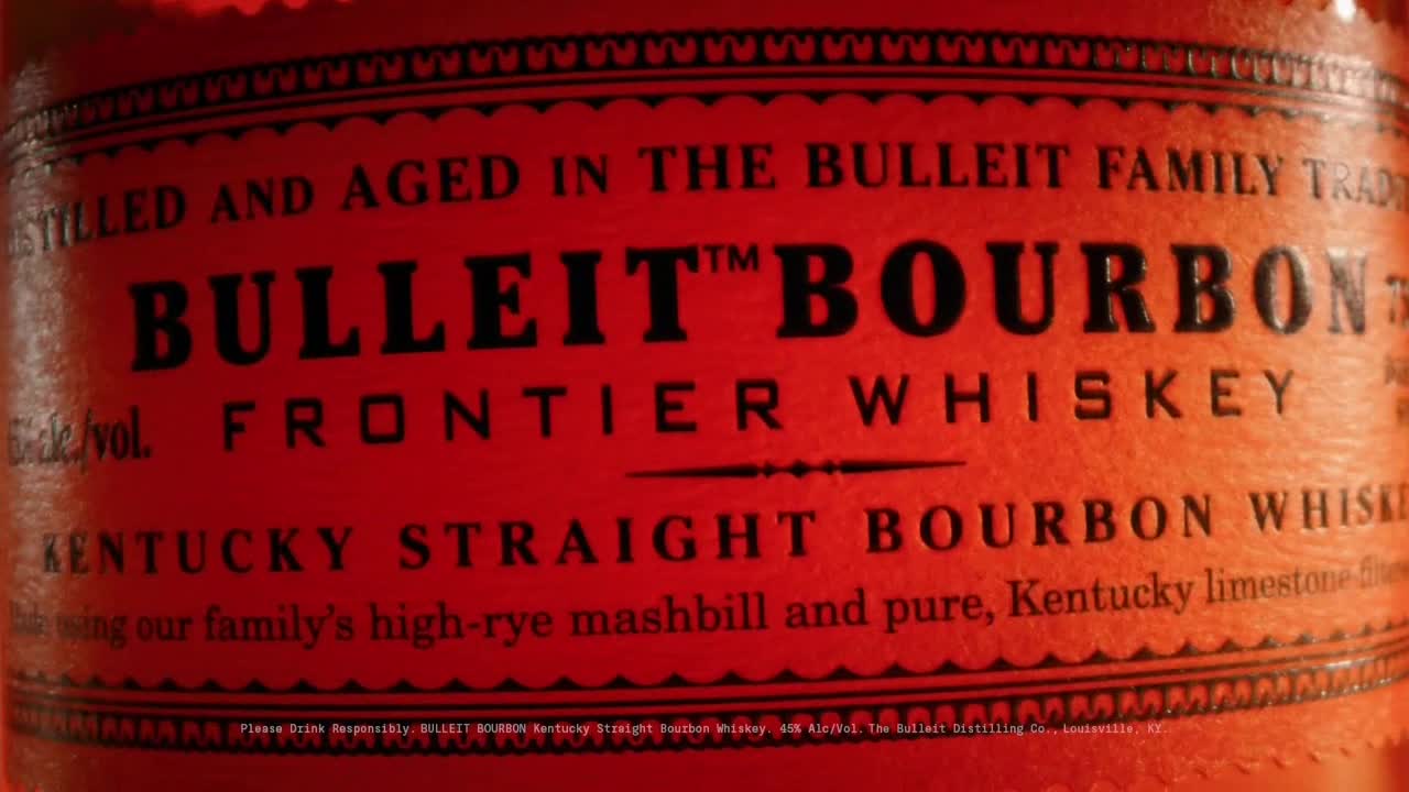 The label of a Bulleit Bourbon bottle is centered, the words "BULLEIT BOURBON" in bold black letters against a reddish-orange background. Below, "FRONTIER WHISKEY" is also visible, and the label is framed with a decorative border.
