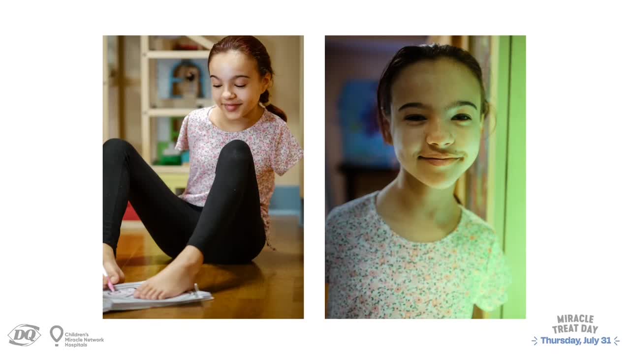 A young girl with a painted-on mustache smiles at the camera, illuminated by green light. Across the frame, she sits cross-legged, focused on a piece of paper in front of her.
