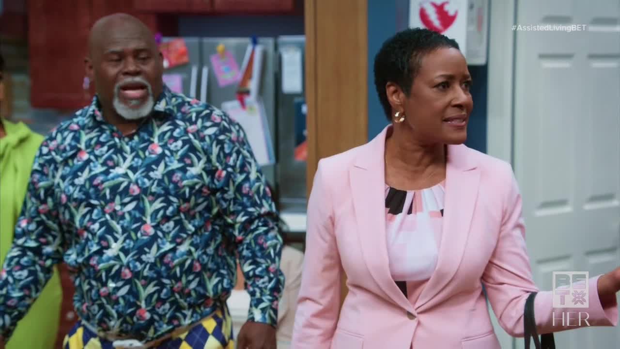 The man in the floral shirt is mid-sentence, mouth open, while the woman in the pink blazer gestures with her hand. They appear to be in a domestic setting, perhaps a scene from a BET Her show.
