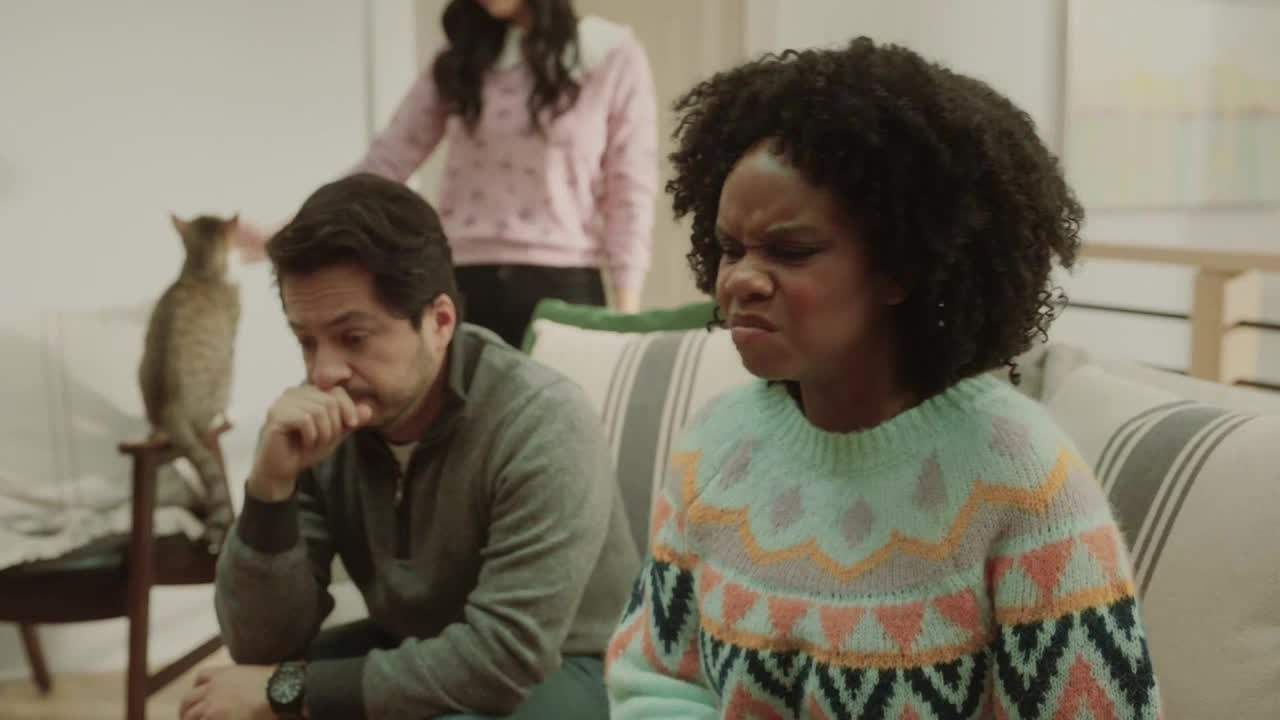 A woman on the couch furrows her brow, clearly displeased. Beside her, a man looks down, his hand covering his mouth, while a cat sits perched on a stool nearby.
