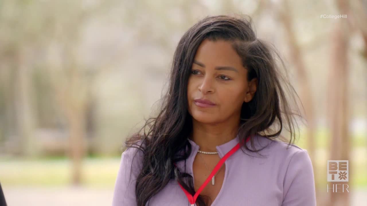 A woman with long, dark hair looks slightly off-camera, her expression thoughtful. She wears a light purple top, a red lanyard around her neck, and a delicate necklace.
