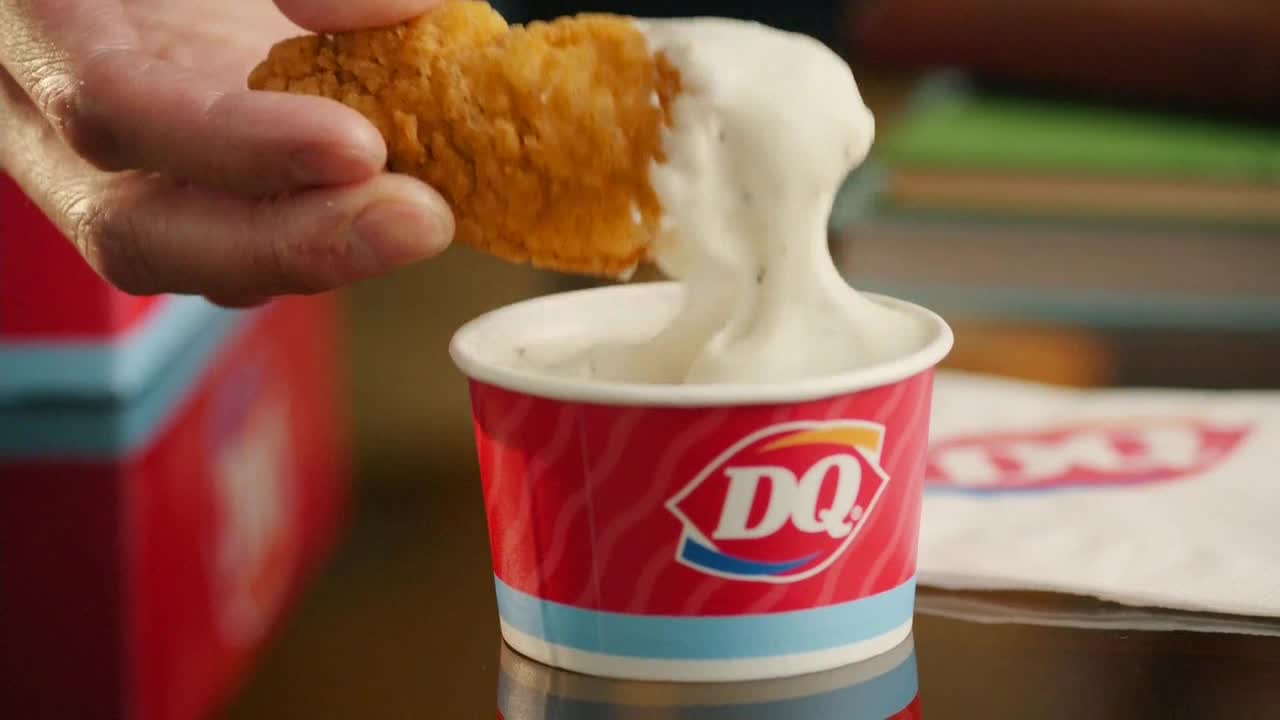 A hand dips a crispy, golden chicken tender into a cup of thick, white ranch dressing. The DQ cup is red and white, and the dressing clings to the chicken as it's lifted.
