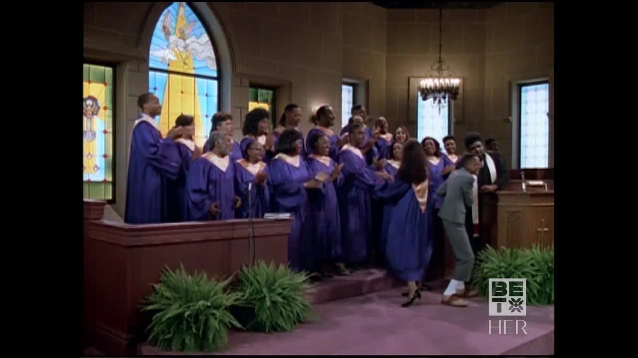 A choir in purple robes claps and smiles as a woman in a robe steps down from the altar. A man in a gray suit follows her, seemingly gesturing.
