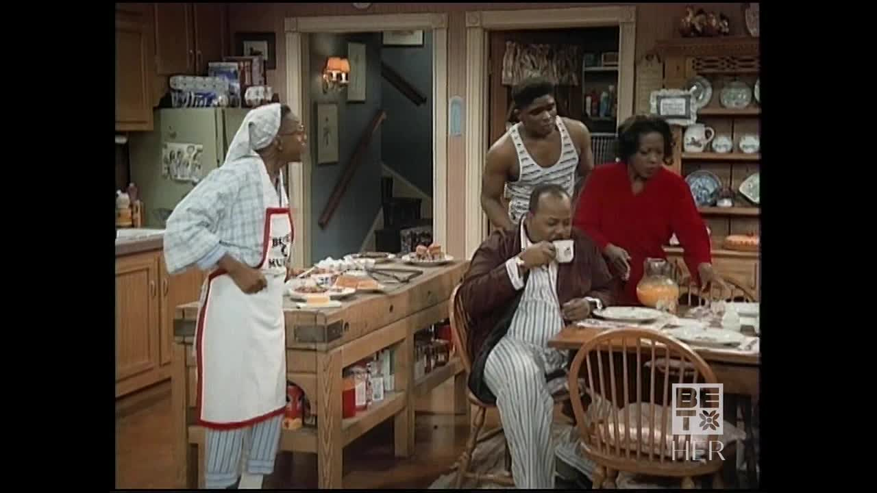 In a cozy kitchen, a woman in a headscarf and apron stands near a butcher block island, while the man at the table sips from a mug. A young man in a tank top hovers behind the seated man, and the woman in red reaches for something on the table.
